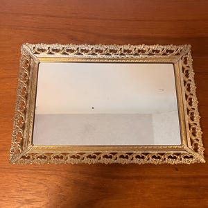 Peut inclure: Un miroir de vanité rectangulaire avec un cadre filigrané doré orné. Le miroir reflète un mur blanc uni. Le cadre présente un design ajouré détaillé sur les bords, ajoutant une esthétique vintage.