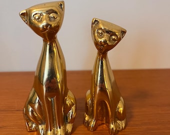 May include: Two gold-colored brass cat figurines. The larger cat is sitting upright with its tail tucked between its legs. The smaller cat is sitting upright with its tail curled around its legs.