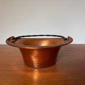 May include: A hammered copper bowl with a wide, flared rim and a black, twisted metal handle attached to two side loops. The bowl features a textured, dimpled surface throughout the exterior and interior, showcasing a warm, metallic orange-brown finish.