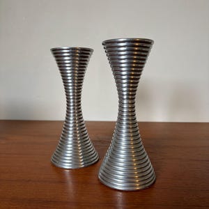 May include: Two silver-colored, spiral-shaped candle holders. The candle holders have a wide base that tapers to a narrow center, then flares out again at the top. They are made of metal and sit on a wooden surface.