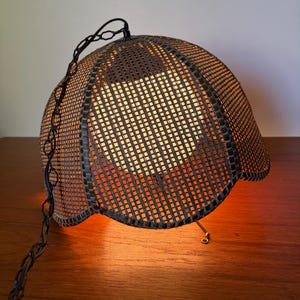 Könnte beinhalten: Eine kuppelförmige Hängelampe mit einem dunkelbraunen, gewebten Netzgeflecht und einer warm leuchtenden Lichtquelle im Inneren. Die Leuchte ist an einer dunklen Metallkette aufgehängt und verfügt über eine kleine Zugkette zur Bedienung.