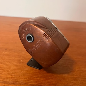 Pode incluir: Um apontador de lápis castanho vintage com a palavra "MIDGET" impressa na frente. O apontador tem uma base retangular e um topo arredondado com um anel de metal prateado em volta do orifício de entrada do lápis. O apontador é feito de um material castanho, possivelmente baquelite.
