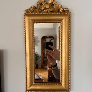 May include: A rectangular mirror with a decorative, ornate frame. The frame is gold-coloured and carved with a rope-like design and a decorative bow at the top.
