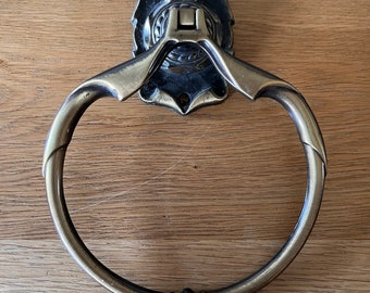 May include: A circular towel ring with a brushed antique brass finish. The mounting plate features a decorative scalloped edge with two screw holes. The ring is attached to a central hinge mechanism that allows for movement, and it includes a small decorative bead detail at the bottom center of the ring.