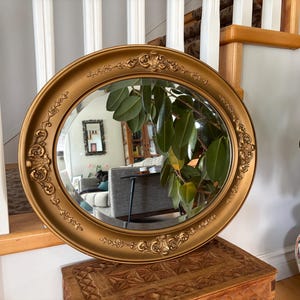 Peut inclure: Un miroir mural ovale doté d'un large cadre doré mat avec des détails ornementaux en relief, incluant des volutes et des motifs floraux sur les bords supérieur et inférieur. La surface en verre réfléchissant montre un intérieur de pièce.