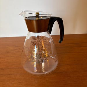 May include: A vintage glass coffee carafe with a brown metal top and black handle. The clear glass body features a gold floral design. The carafe is designed with a pouring spout and a gold-coloured lid.