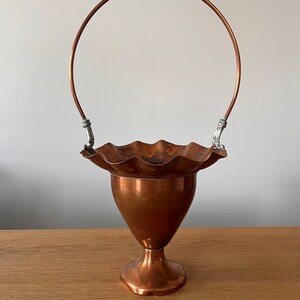 May include: A copper vase with a fluted rim and a handle. The vase has a pedestal base and is made of polished copper.