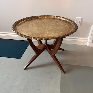 Puede incluir: Una mesa de bandeja redonda de latón antiguo con un borde decorativo festoneado. La bandeja se asienta sobre una base de madera oscura con un diseño de patas en forma de X. La mesa tiene unos 61 cm de diámetro.