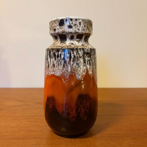 Puede incluir: Un jarrón de cerámica con un diseño único. La parte superior está moteada con marrón y negro, pasando a un patrón de goteo blanco y negro. La sección media es naranja y la base es marrón oscuro. El jarrón está sobre una superficie de madera.