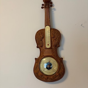 May include: A brown violin-shaped wall instrument featuring an integrated thermometer with Fahrenheit and Celsius scales and a circular barometer dial with weather indicators including Stormy, Rain, Change, Fair, and Very Dry printed on the face.