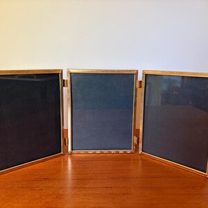 Könnte beinhalten: Ein Triptychon-Bilderrahmen mit drei verbundenen rechteckigen Paneelen in metallischem Gold-Finish. Jeder Rahmen hat einen strukturierten, dekorativen Rand und enthält einen dunklen, einfarbigen Einsatz. Die Paneele sind durch Scharniere verbunden.