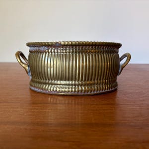 May include: A round brass bowl featuring a vertical ribbed texture along the exterior walls, a beaded rim, and two small curved handles on opposite sides. The metal surface displays a warm, metallic gold tone with signs of natural patina.