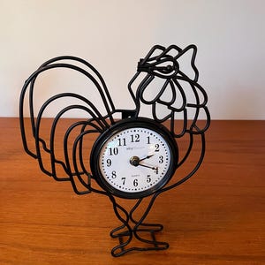 Op de afbeelding: Een zwarte metalen haanvormige klok met een witte wijzerplaat. De wijzerplaat heeft zwarte cijfers en wijzers. De klok is gemaakt van een draadachtig materiaal en staat op een houten oppervlak. De klok is een decoratief item.