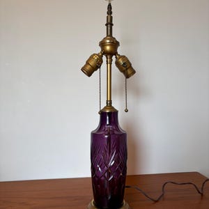 May include: A vintage table lamp with a deep purple glass base featuring a diamond-cut pattern. The lamp has a brass-colored metal base and a three-bulb fixture. The lamp is topped with a small, clear glass shade and a decorative finial.