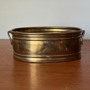 Puede incluir: Un cuenco ovalado de latón con un acabado metálico liso y pulido, que cuenta con dos pequeñas asas curvas fijadas a los lados. El recipiente tiene un borde redondeado sencillo y una banda horizontal continua en la pared exterior.