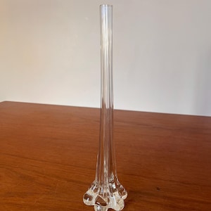 May include: A clear glass bud vase with a long, slender neck and a flared, footed base. The vase has vertical ridges along its length and sits on a wooden surface. The vase is approximately 30 cm tall.