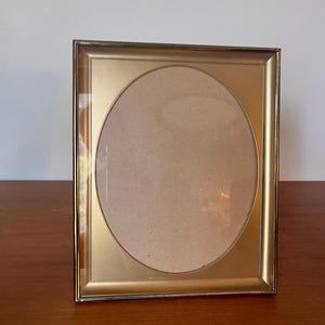 May include: A rectangular gold-colored picture frame with an oval opening. The frame has a glass front and a cardboard backing. The frame is sitting on a wooden surface.