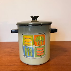 Może przedstawiać: Szary garnek emaliowany z czarnymi uchwytami i czarną pokrywką. Garnek ma graficzny wzór czterech kwadratów z różnymi warzywami i kiełbasami w każdym kwadracie.