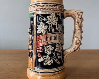 Op de afbeelding: Een keramische bierkan met een bruin, blauw en groen ontwerp. De kan heeft een handvat met een wijnstokontwerp en de tekst "Froh und Heiter" aan de voorkant.
