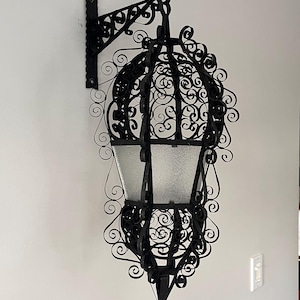 May include: Black wrought iron outdoor hanging lantern with a decorative scroll design. The lantern has a clear glass enclosure and is mounted on a white wall. The fixture is designed to hold a light source.