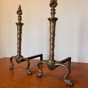 Puede incluir: Dos morillos antiguos de latón. Cada uno presenta una parte superior en forma de llama, una columna detallada con un diseño de serpiente y patas curvas con garras. Los morillos están sobre una base de metal negro.