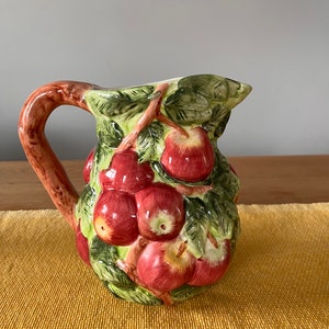 Puede incluir: Una jarra de cerámica con asa marrón y un diseño de manzanas rojas y hojas verdes.