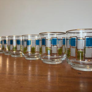 May include: A set of eight vintage lowball glasses. Each glass has a gold rim and a geometric pattern of teal, white, and green rectangles. The glasses are arranged in a row on a wooden surface.