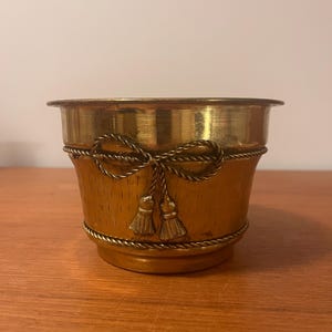 May include: A small, gold-colored metal planter with a textured, light brown exterior. The planter is adorned with a decorative rope and tassel design. The rim and base are also gold-colored.