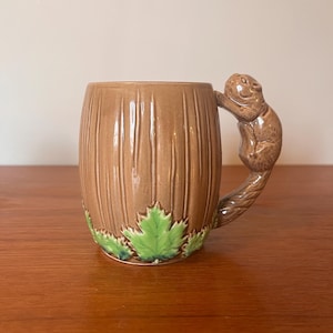 Puede incluir: Una taza de cerámica marrón con un asa de ardilla y detalles de hojas verdes. La taza tiene forma de tronco de árbol.