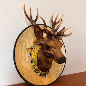 Puede incluir: Una escultura decorativa de cabeza de ciervo montada sobre una placa redonda de madera con un borde negro. La cabeza de ciervo es marrón con cuernos detallados y está colocada contra un elemento decorativo amarillo y blanco.
