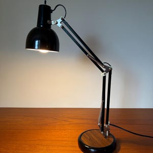 May include: A black adjustable desk lamp with a dome-shaped shade and a circular base featuring a wood grain finish. The lamp has a multi-jointed metal arm with silver-toned hardware and a black power cord extending from the base.