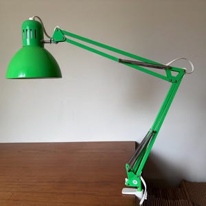 May include: A bright green architect desk lamp with an adjustable articulated arm and a dome-shaped shade. The lamp features a white power cord and a white clamp base designed to attach to the edge of a flat surface for focused task lighting.
