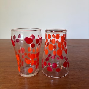 Könnte beinhalten: Zwei klare Trinkgläser, die mit einem Muster aus leuchtend roten und orangefarbenen Punkten unterschiedlicher Größe verziert sind. Ein Glas steht aufrecht, während das andere umgedreht ist und die kreisförmigen Motive auf der Außenfläche zeigt.