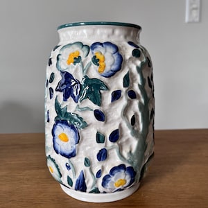 May include: A white ceramic vase with a raised floral design. The flowers are blue, yellow, and green. The vase has a green rim.