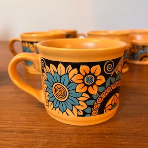 May include: Four ceramic mugs with a yellow exterior and a black band featuring a floral design in teal and orange. Each mug has a curved handle. The mugs are on a wooden surface.