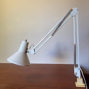 May include: A white desk lamp with an adjustable articulated arm, a conical shade, and exposed metal springs. The lamp features a clamp-style base for attachment to a surface and includes a white power cord extending from the back of the light fixture.