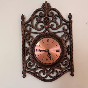 May include: Ornate wall clock with a copper-colored face and black Roman numerals. The clock is encased in a decorative, dark bronze-colored metal frame with scrolling details and a floral accent at the top. The hands point to approximately 5:40.