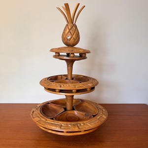 May include: A three-tiered wooden serving tray with a pineapple-shaped top. The tray has carved details and circular levels with divided sections for snacks or appetizers. The wood has a warm, natural finish.