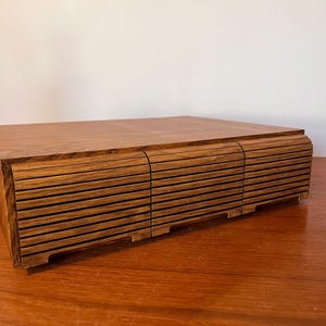May include: A rectangular wooden storage box with a smooth top surface and three front-facing drawers. Each drawer features a horizontal slatted design with a curved profile, creating a textured appearance across the front of the unit.
