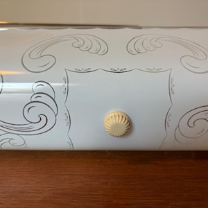 May include: A white rectangular light fixture cover featuring a silver scrollwork pattern and a central cream-colored circular knob. The surface is smooth and glossy, with a scalloped edge along the top and a clean, symmetrical design.