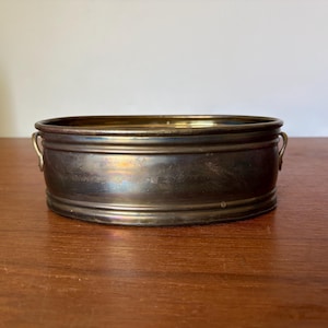 May include: An oval-shaped brass bowl with a metallic finish and a dark, aged patina. The bowl features a smooth, rounded rim, a slightly ribbed body, and two small, curved handles attached to the sides, providing a simple and functional design.