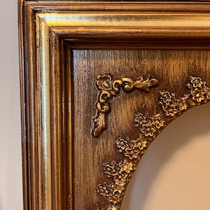 Może przedstawiać: Ozdobna, złota rama do zdjęć z bogatą, teksturowaną powierzchnią. Rama posiada dekoracyjne akcenty kwiatowe i wici, a także warstwowy design. Rama ma estetykę vintage.