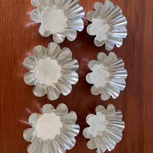 May include: Six silver-colored, fluted, round baking cups with a white powder inside, arranged on a brown wooden surface.