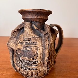 May include: A brown ceramic pitcher with a handle and a flared rim. The pitcher features an embossed design with the text "1928 Ford Models" and a car illustration. The design is wrapped around the body of the pitcher.