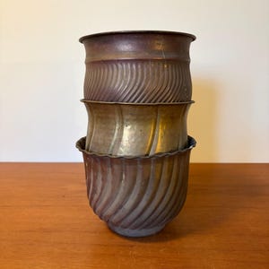 Könnte beinhalten: Drei antike Metallpflanzgefäße, die auf einer Holzoberfläche gestapelt sind. Die Pflanzgefäße haben ein geriffeltes, spiralförmiges Design und eine verwitterte, bronzeähnliche Oberfläche. Der obere Pflanztopf hat einen dunkleren Bronzeton.