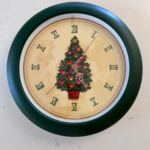 May include: A round wall clock with a dark green frame. The clock face features a Christmas tree with ornaments and a red basket. Roman numerals mark the hours. The clock hands are red and gold.