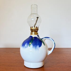 May include: A white ceramic oil lamp with a round base and handle, adorned with blue brushstrokes. It features a clear glass globe and chimney, with a brass fitting. The lamp is designed for decorative or functional use.