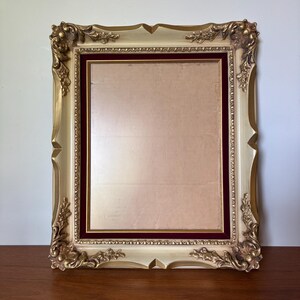 May include: A rectangular picture frame featuring a cream-colored finish with gold-toned accents. The frame includes ornate corner detailing and a deep burgundy velvet inner border surrounding a central rectangular opening for a photograph.