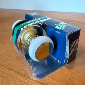 May include: A new door handle set with a gold finish and a white and blue handle. The product is in its original packaging, which is blue and white and has the brand name "WEISER" printed on it.