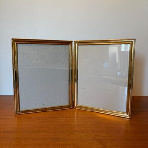 以下が含まれることがあります： 金色の金属フレームが付いた、蝶番式の二連フォトフレーム。 フレームは空で、薄い灰色の裏地が付いています。 フォトフレームは開いており、木製の表面に置かれています。
